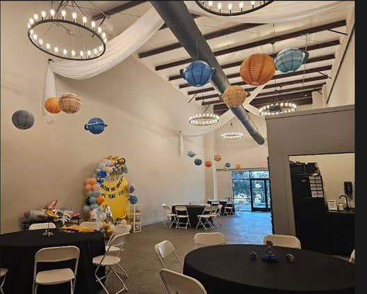 The room set for a party, with round tables, paper lanterns hanging from the ring pendants and a balloon arch at the far wall