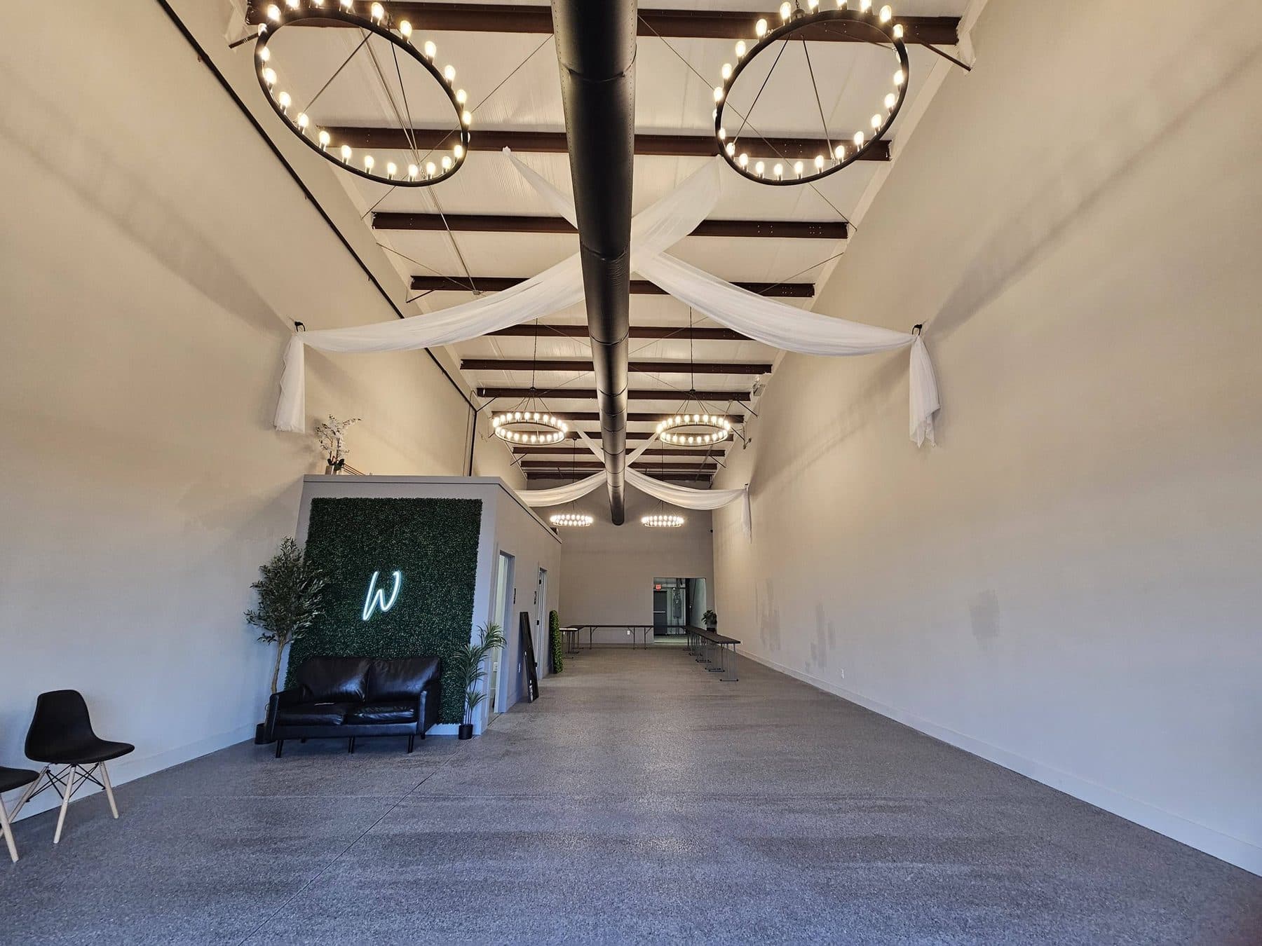 The Willow Tree Events room: polished floors running past black chairs to a boxwood wall carrying a white W monogram, lit by circular ring pendants under a black ceiling
